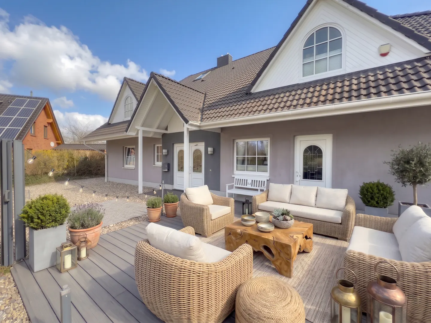 Outdoor patio with wicker furniture, wood table, and potted plants. Gray house with white trim and brown roof in the background.