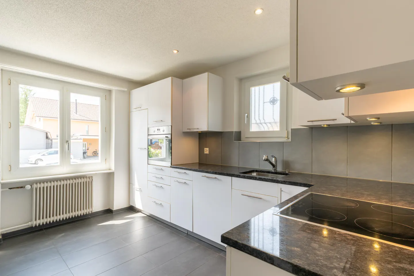 Bright kitchen with white cabinets, dark countertops, and gray tile flooring. A window overlooks a residential street.