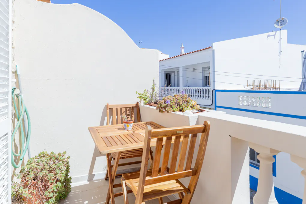 A sunny balcony with a wooden table and chairs, potted plants, and a view of white buildings under a clear blue sky.