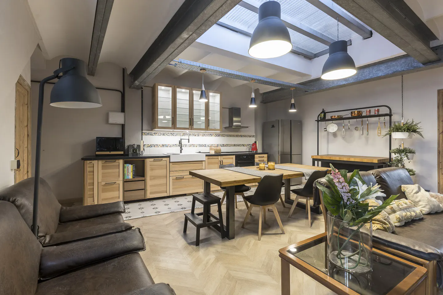 Open-concept living space with wood floors, exposed beams, and skylights. Kitchen with light wood cabinets and stainless steel appliances. Brown leather sofas.