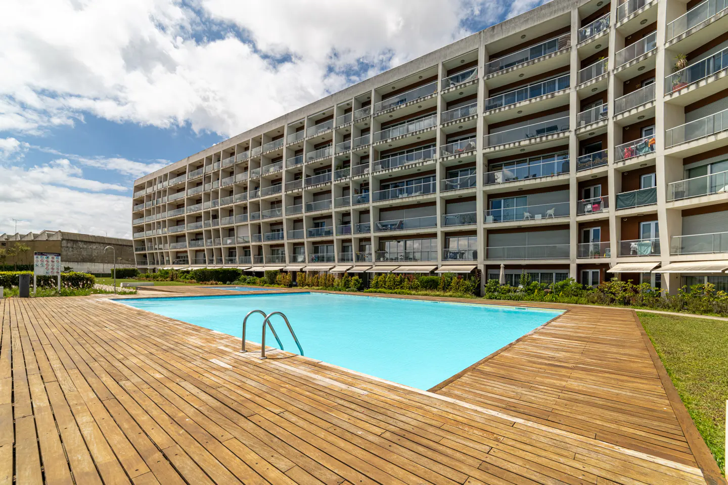 Outdoor pool with wooden deck and metal ladder in front of a large white apartment building under a cloudy sky.