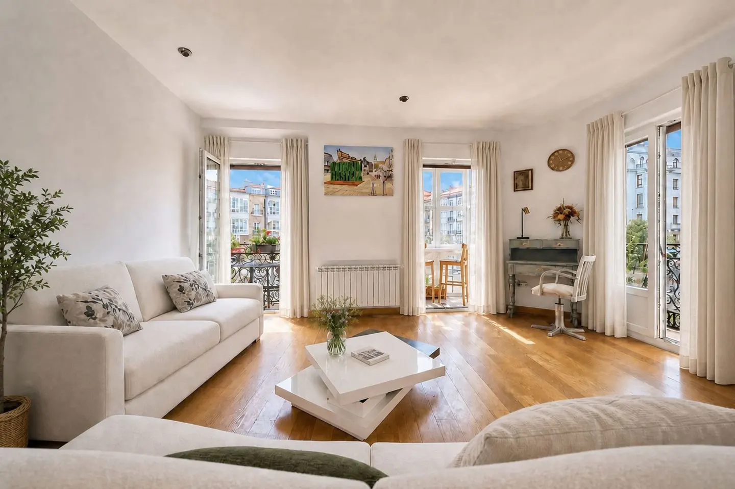Bright living room with white sofas, wood floors, and balconies. A modern white coffee table sits in the center. Natural light streams in.