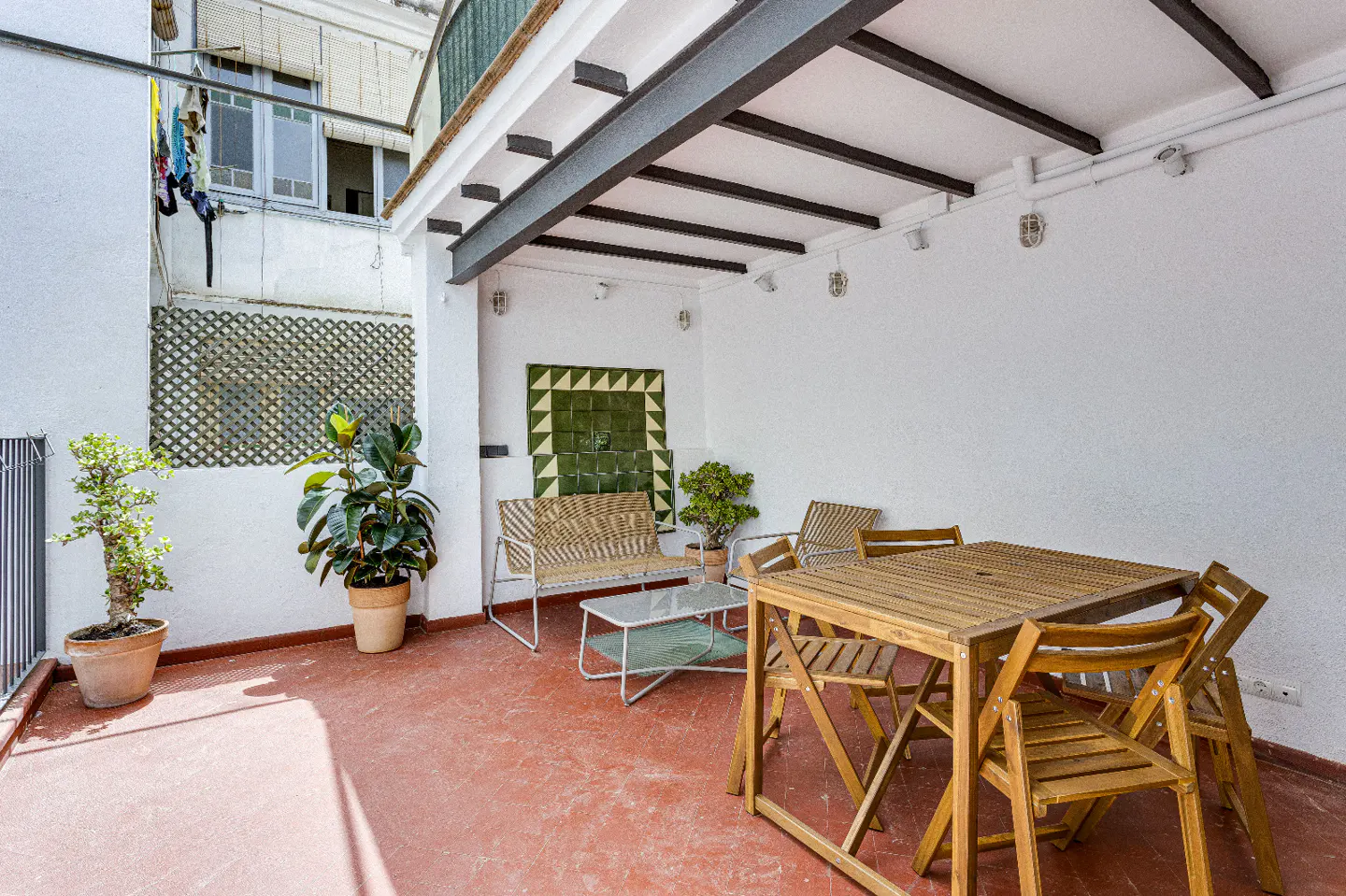 Outdoor patio with a wooden table and chairs, woven benches, potted plants, and a red tile floor.