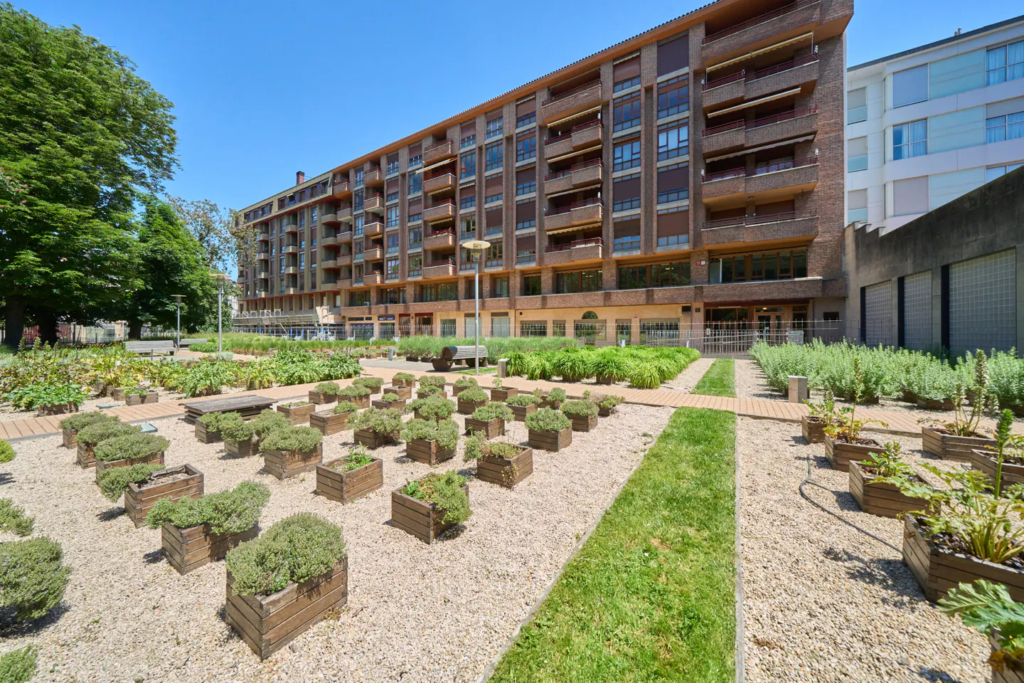 Brick apartment building with balconies overlooks a garden with wooden planters and gravel paths on a sunny day.