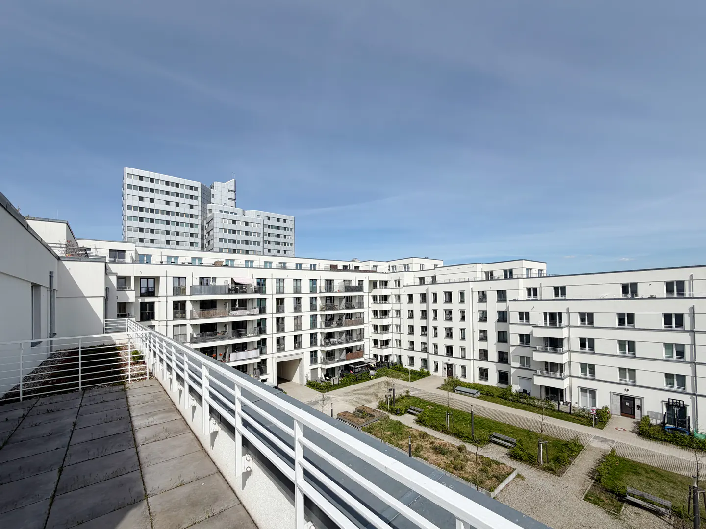View of a modern, white apartment complex with balconies surrounding a courtyard with benches and greenery under a blue sky.