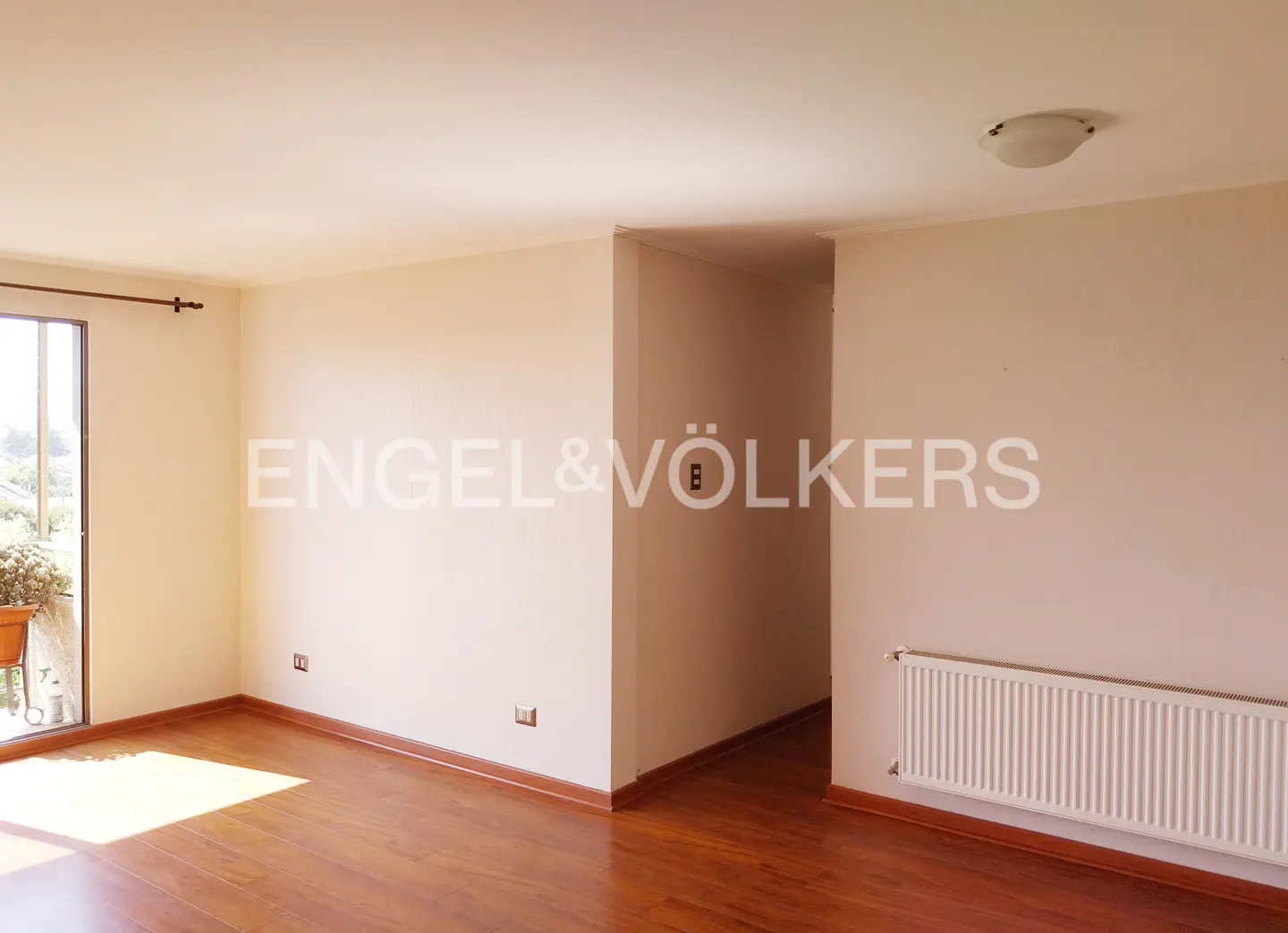 An empty room with hardwood floors, beige walls, and a balcony door. A radiator is on the wall. Engel & Völkers logo is in the center.
