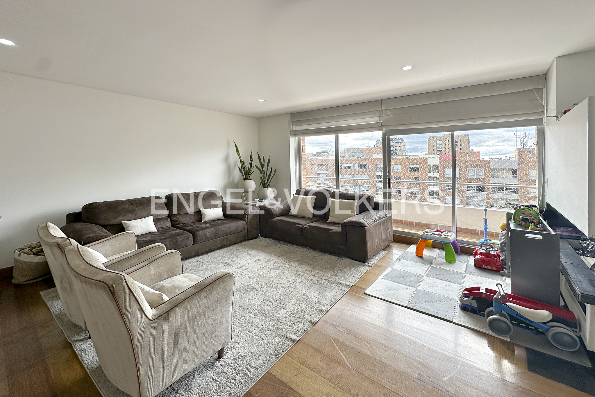 Living room with brown sofas, beige chairs, and a light gray rug. Toys are on a gray and white play mat near a large window.