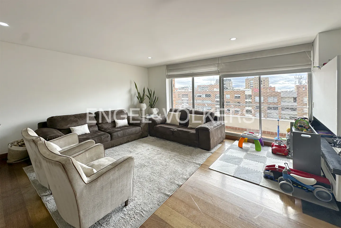 Living room with brown sofas, beige chairs, and a light gray rug. Toys are on a gray and white play mat near a large window.