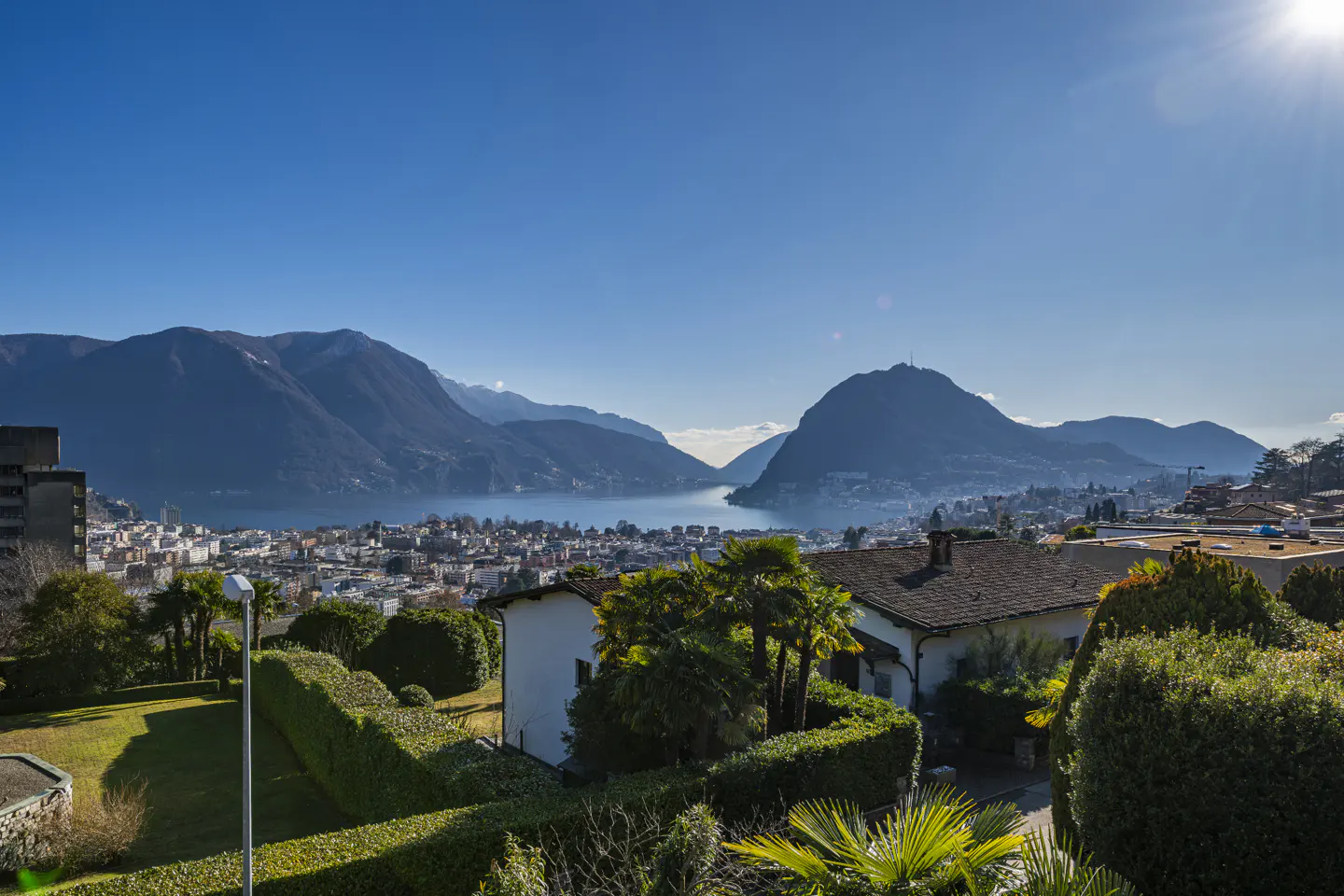 Scenic view of Lugano, Switzerland, with mountains, lake, city, and houses under a clear blue sky.