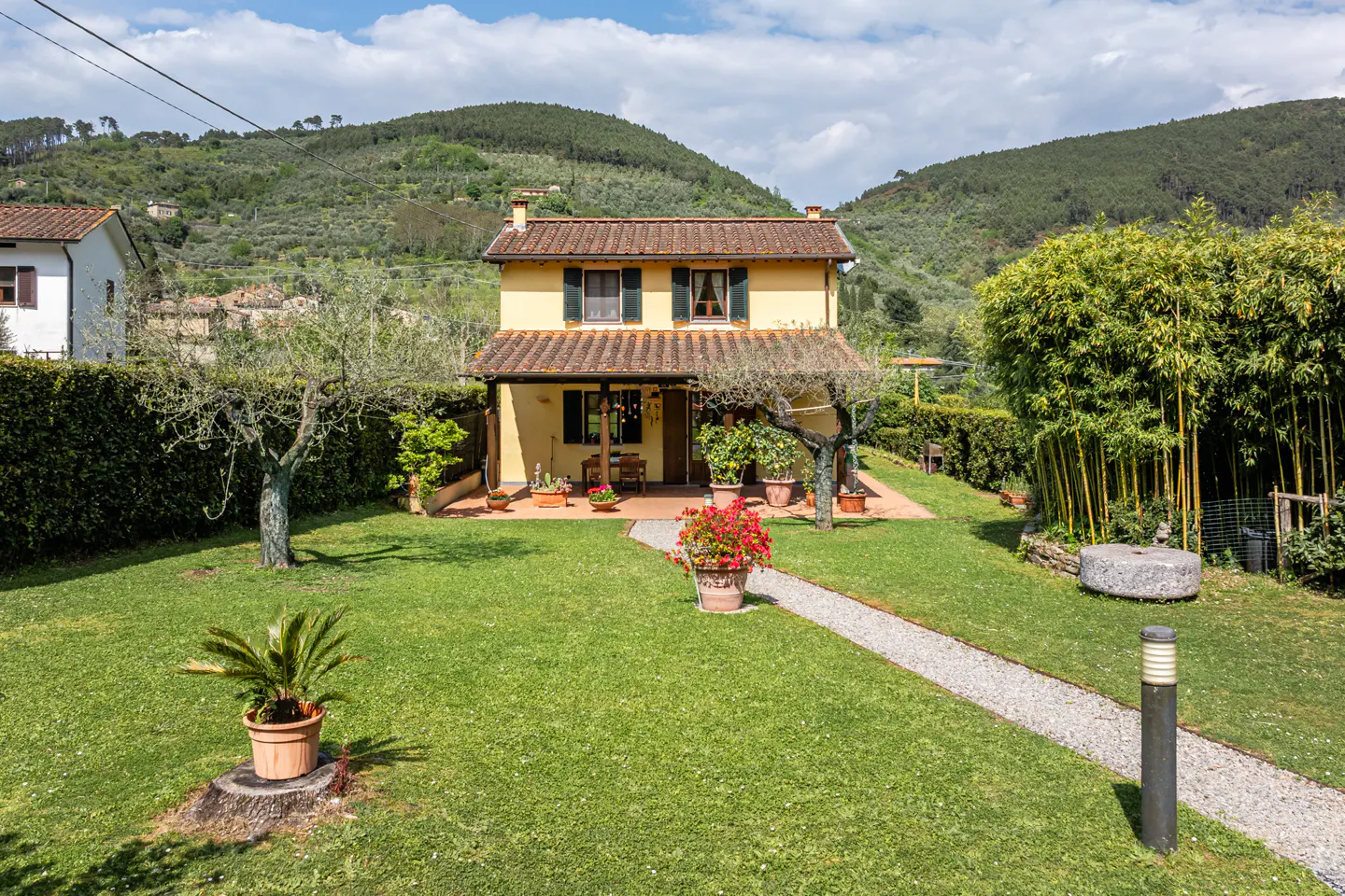 Two-story yellow house with a brown tile roof, green lawn, and stone path leading to the front porch. Rolling green hills in the background.