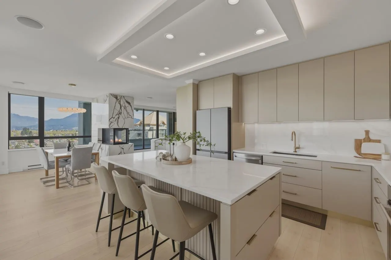 Bright, modern kitchen with a white island, beige cabinets, and bar stools. A marble fireplace and dining area are visible in the background.