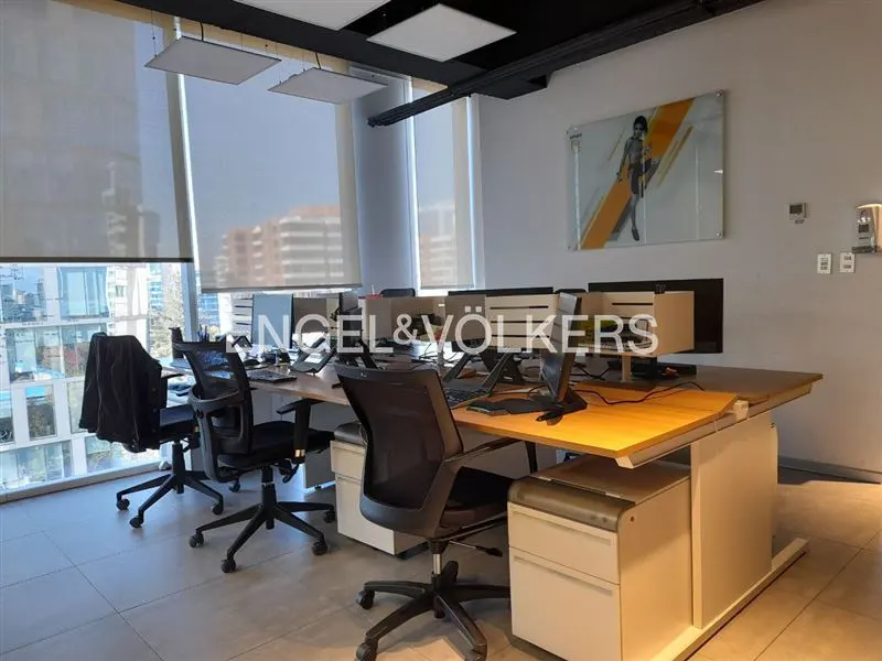 Office space with desks, black chairs, and computers. Large windows with blinds offer a city view. Artwork hangs on the wall.