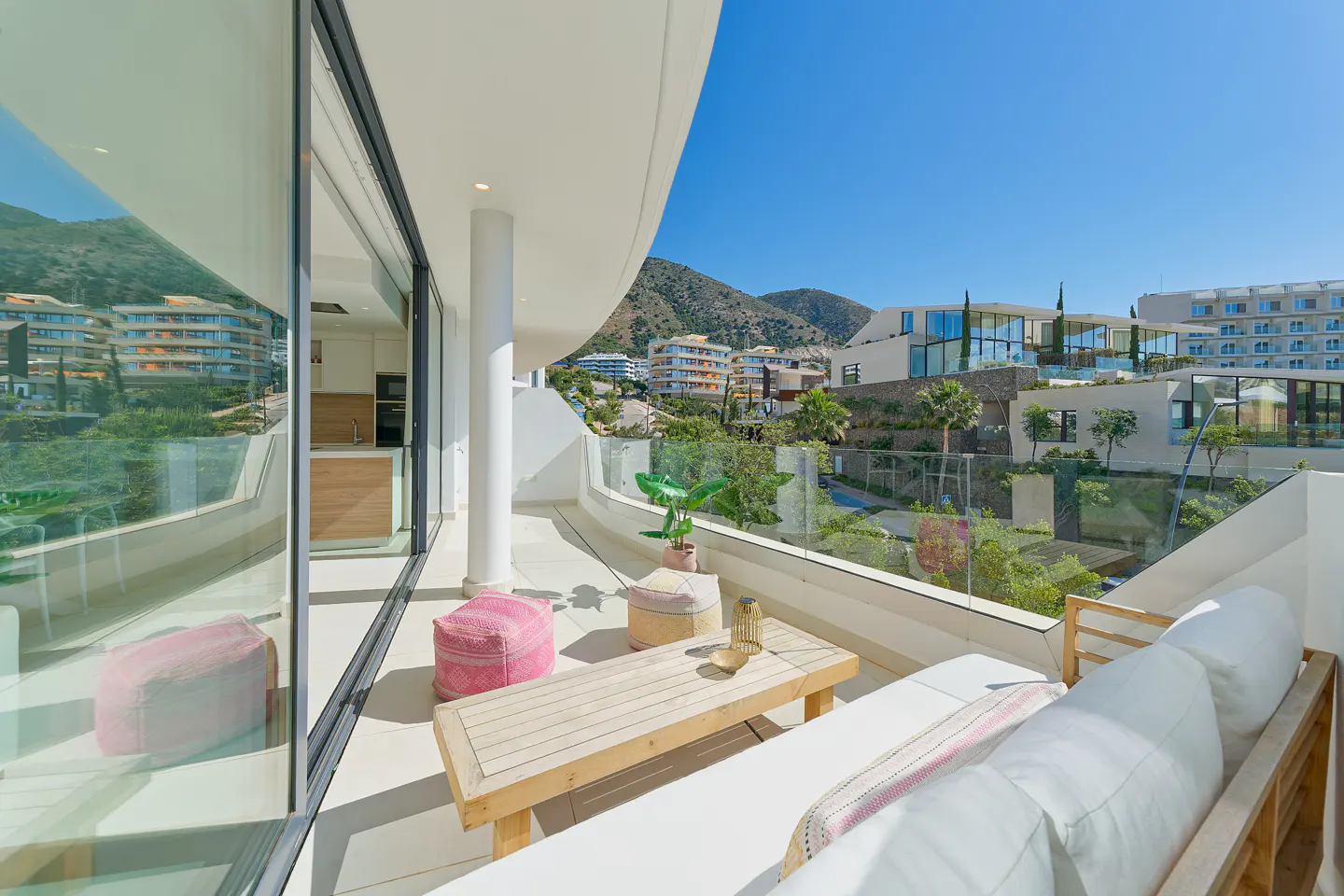 Bright, modern balcony with white lounge seating, pink poufs, and a wooden table. A clear blue sky and mountain views are visible.
