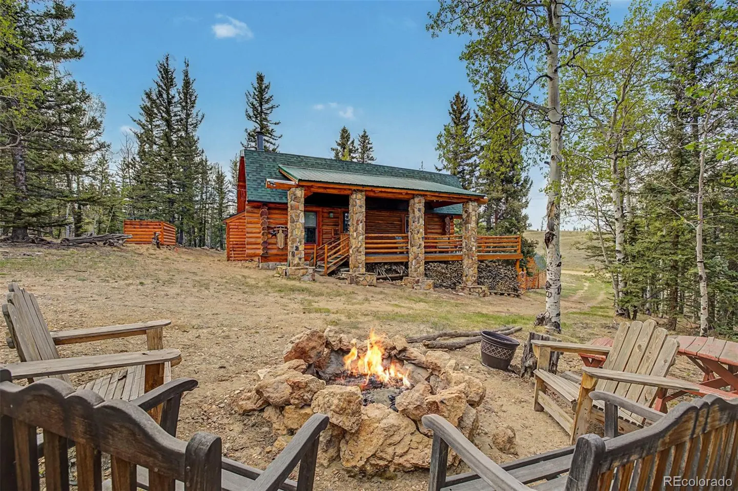 Log cabin with a green roof and stone pillars, surrounded by trees. A fire pit with Adirondack chairs sits in the foreground.