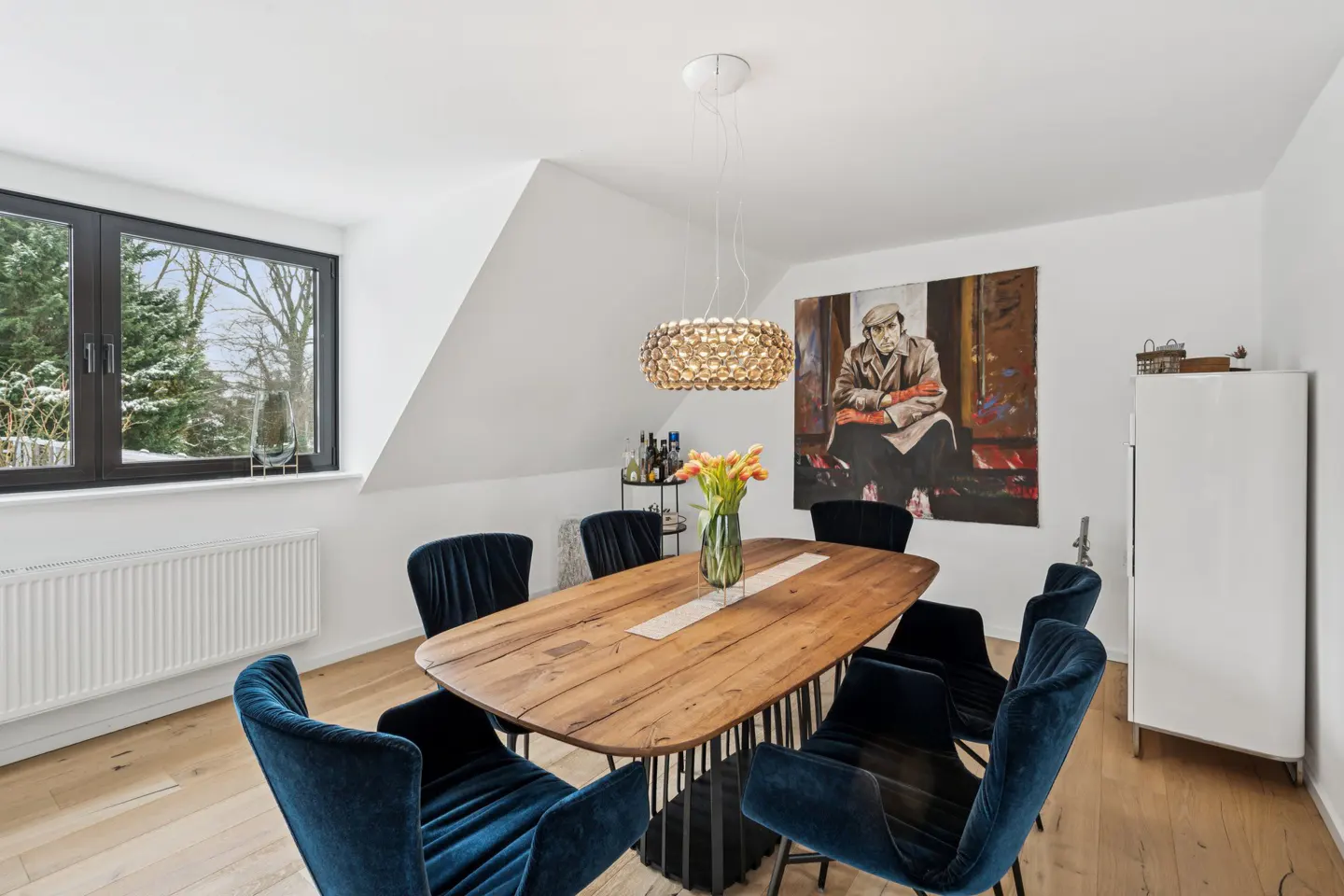 Bright dining room with a wood table, blue velvet chairs, and a modern chandelier. A large painting hangs on the white wall.