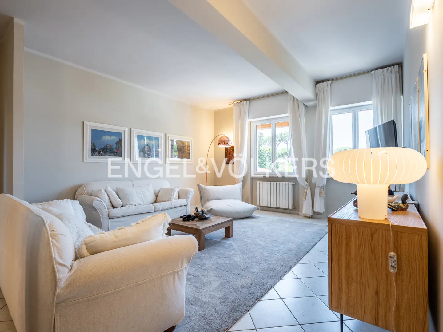 A bright living room with white sofas, a wooden coffee table, and a large gray rug. A mushroom lamp sits on a wooden cabinet. Windows with white curtains let in natural light.
