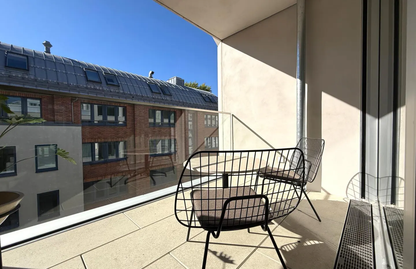 Balcony view with two black wire chairs and a small table. Outside, a brick building with a gray roof is visible under a clear blue sky.
