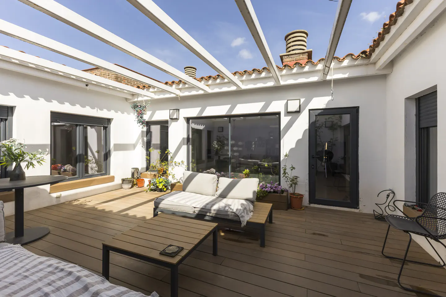 Outdoor patio with wood deck, white walls, and pergola. Furniture includes a sofa, table, and chairs. Plants add a touch of greenery.