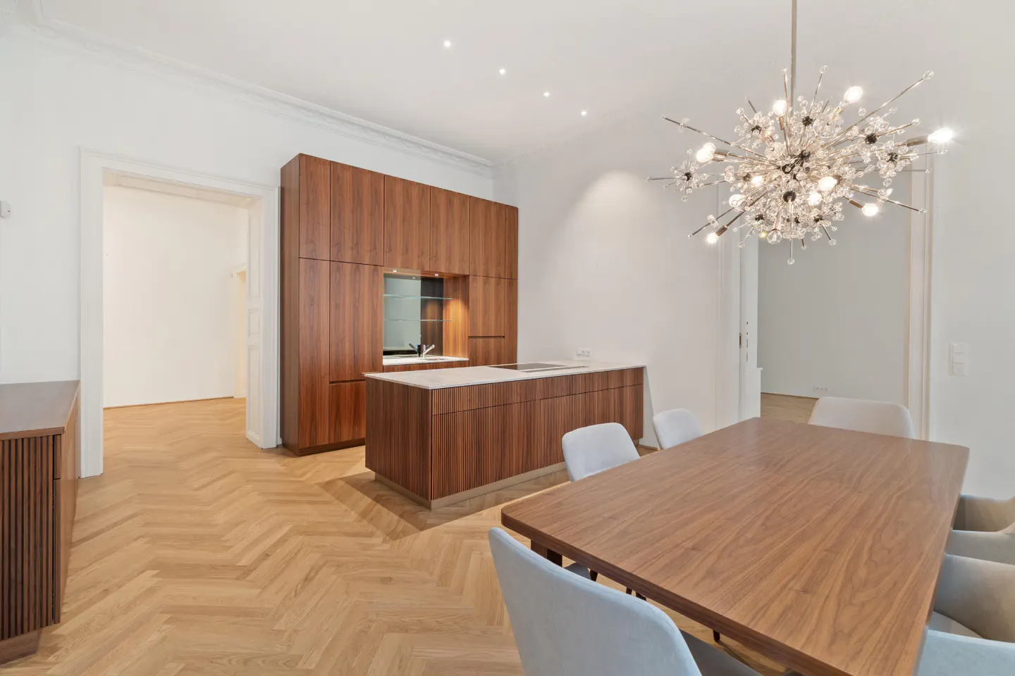 A bright, modern kitchen and dining area with wood floors, cabinets, and a large table with light gray chairs. A crystal chandelier hangs above.