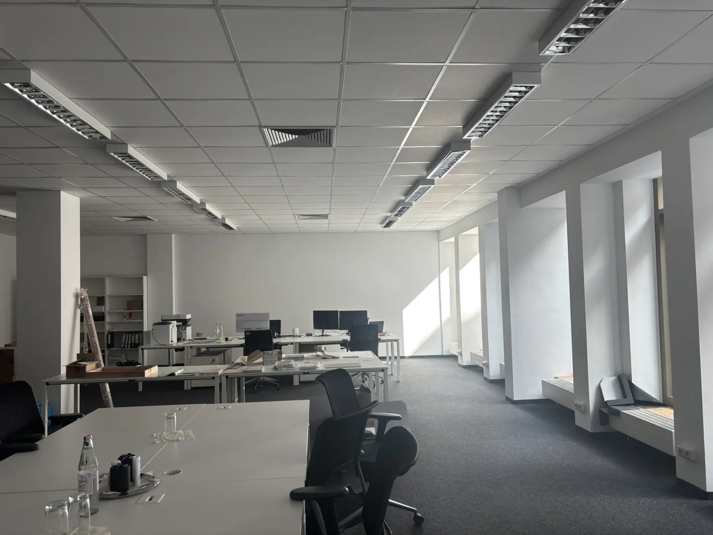 Bright, empty office space with white walls, desks, and chairs. Overhead lighting illuminates the room, and sunlight streams through the windows.