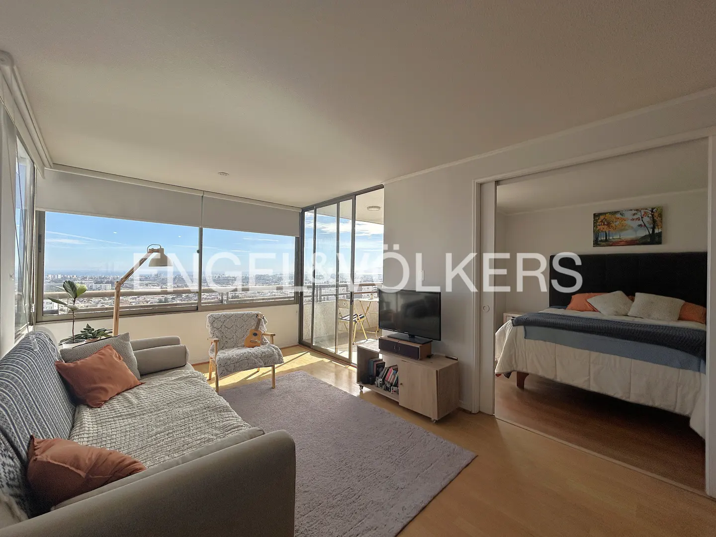 Living room with a gray sofa, chair, TV, and a view of the city. Bedroom with a bed is visible through a doorway.