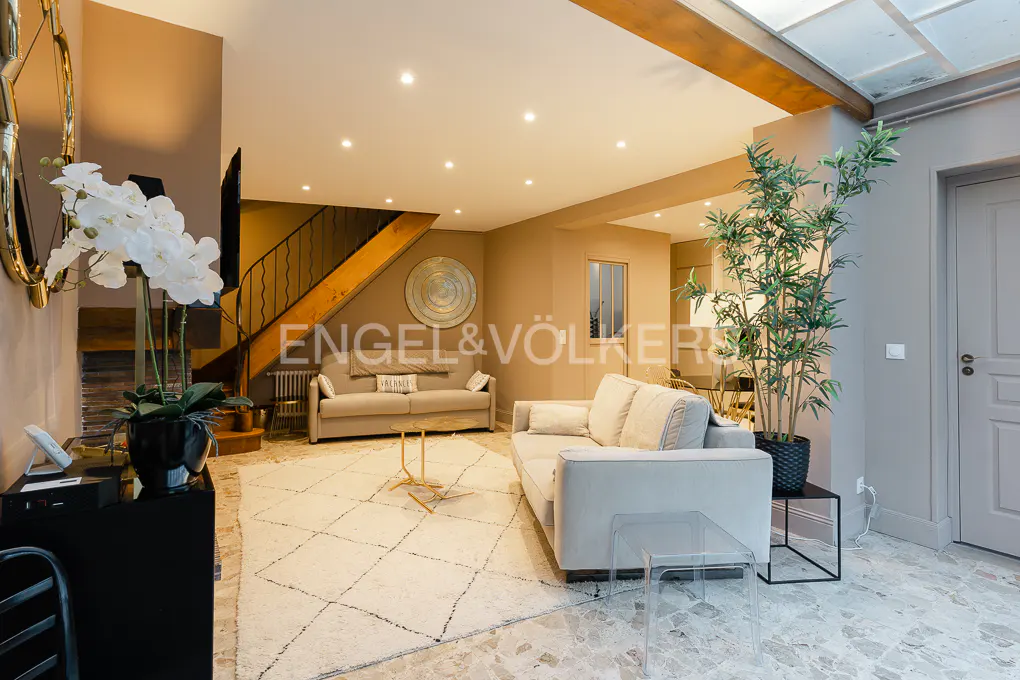 Living room with beige sofas, a white patterned rug, and a staircase in the background. A plant stands near a gray door.