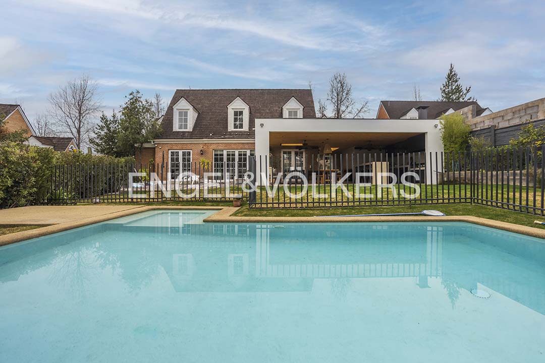 Backyard view of a brick house with a swimming pool, covered patio, and a black metal fence under a blue sky. [AI-generated text]