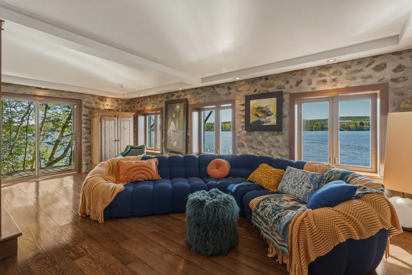 Living room with stone walls, wood floors, and a blue sectional sofa with colorful pillows. Windows overlook a lake.