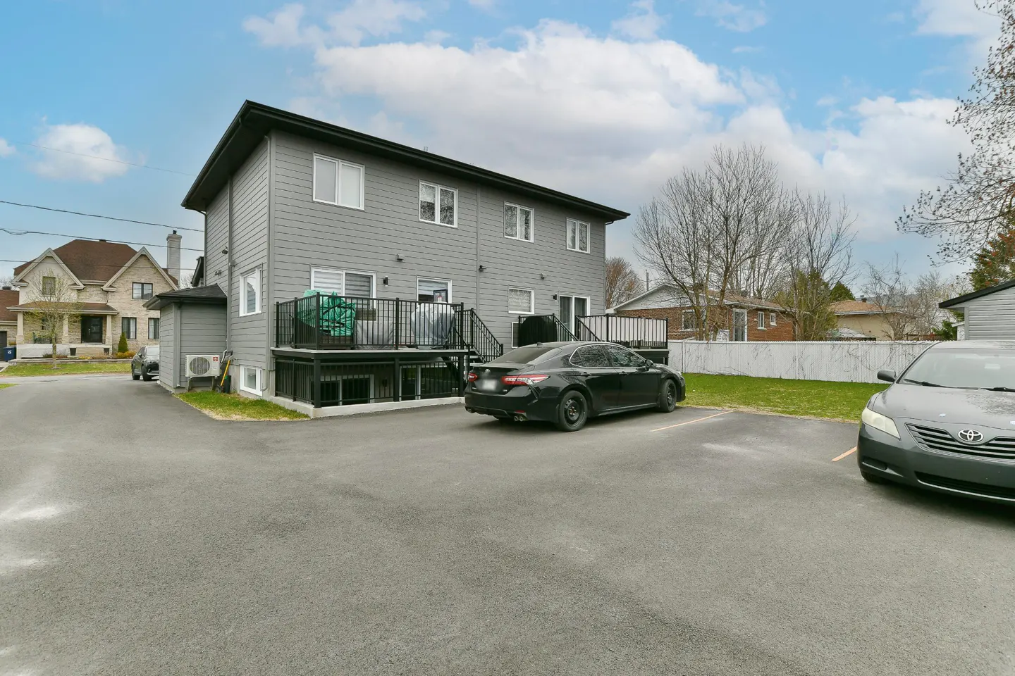 Back of a two-story gray apartment building with a black deck and parked cars in the asphalt parking lot.
