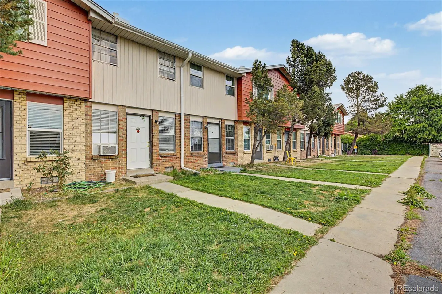 A row of two-story townhouses with brick and siding exteriors, green lawns, and concrete walkways under a blue sky.