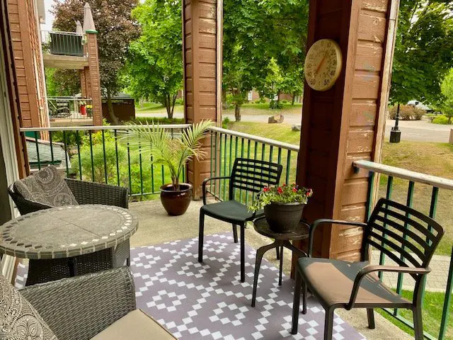 A cozy balcony with wicker furniture, a patterned rug, and potted plants overlooks a green landscape. A thermometer hangs on a brown wooden post.