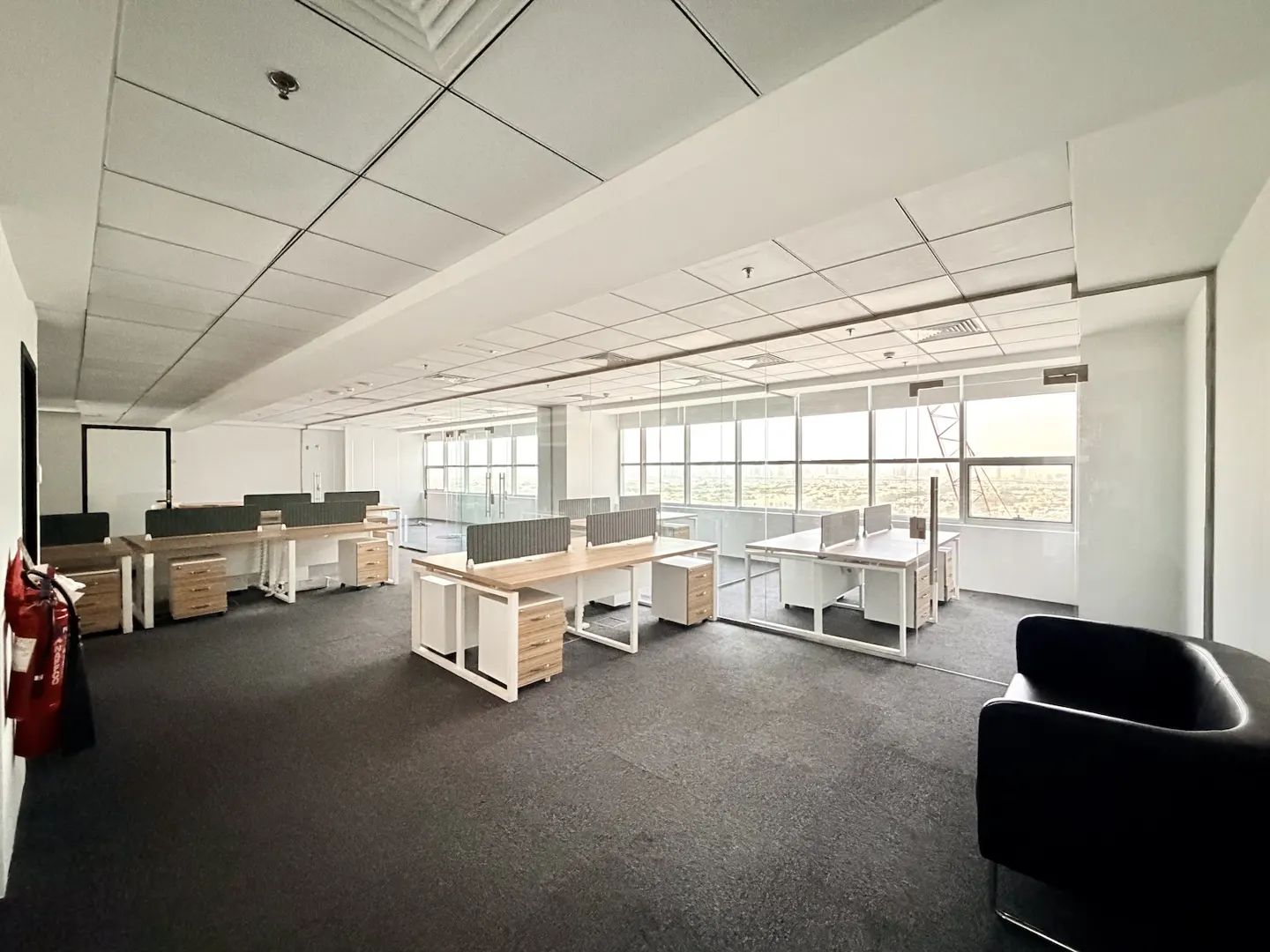 Bright, empty office space with desks, chairs, and large windows. A black couch sits in the foreground.