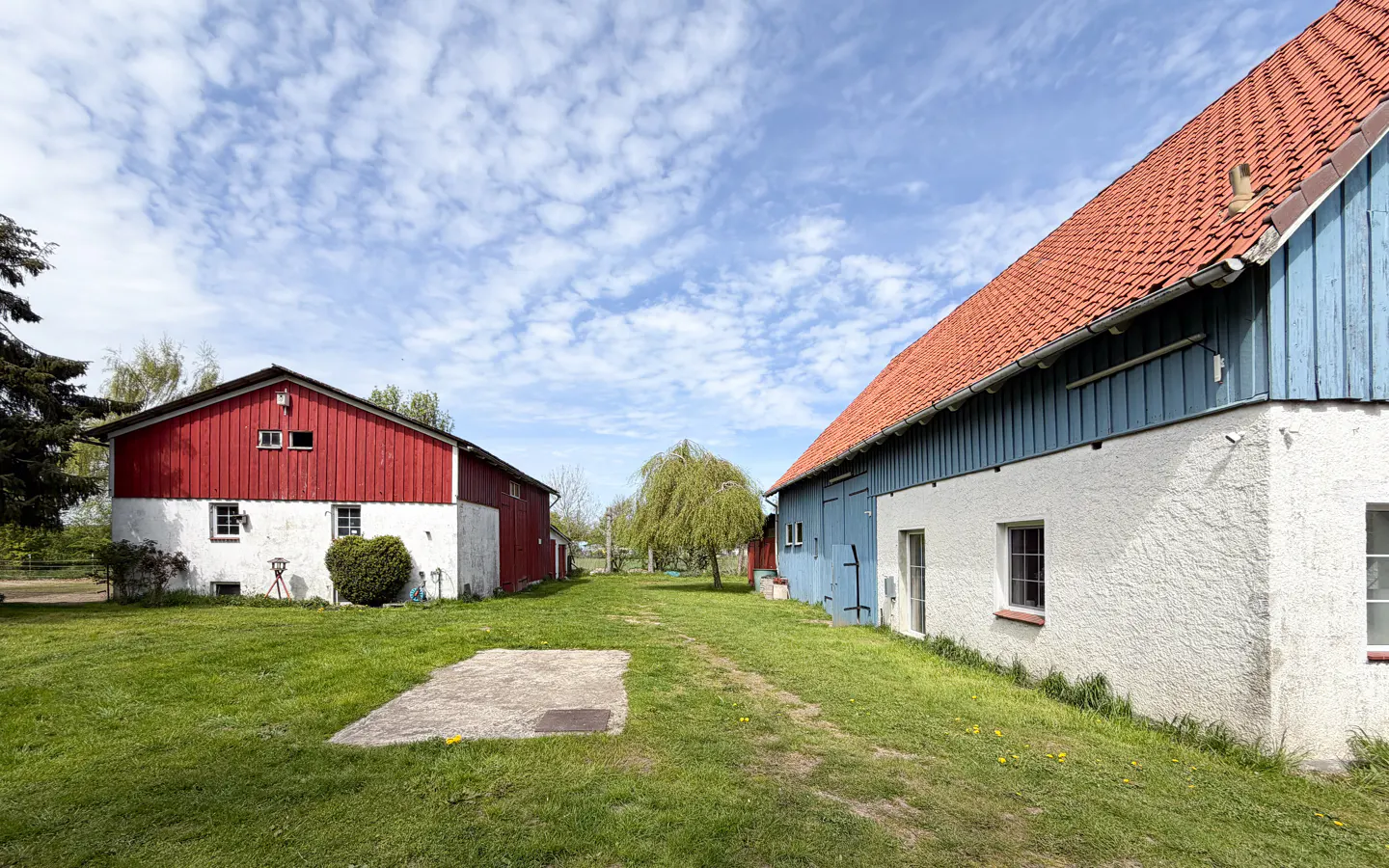 Exterior view of two barns, one red and one blue, on a grassy lawn under a partly cloudy sky.