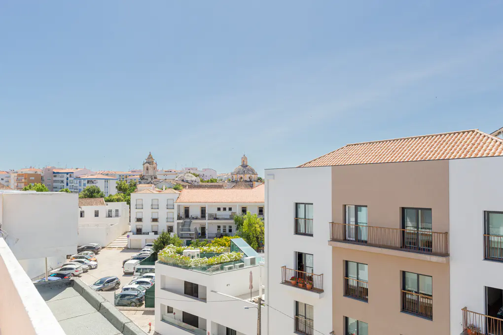 View from a balcony overlooking a sunny Mediterranean town with white buildings, a parking lot, and distant church domes.