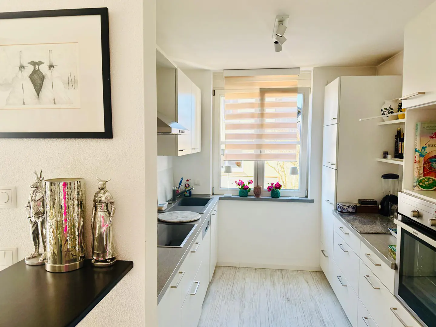 Bright, narrow kitchen with white cabinets, gray countertops, and wood-look floors. A window with blinds sits above the sink. Artwork and figurines decorate the space.