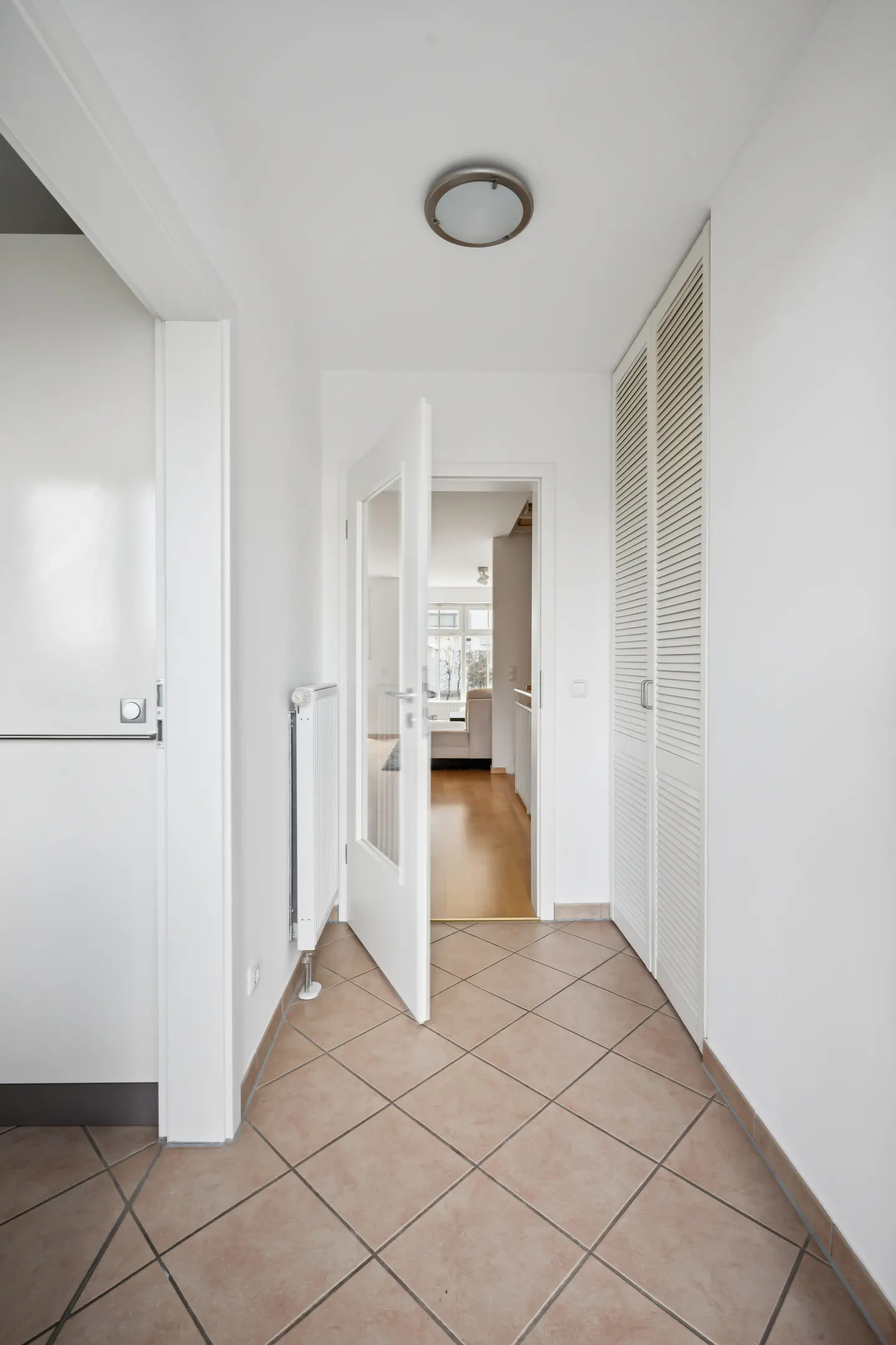 Bright hallway with beige tile floor, white walls, and an open door leading to a sunlit room. A radiator and closet doors are visible.
