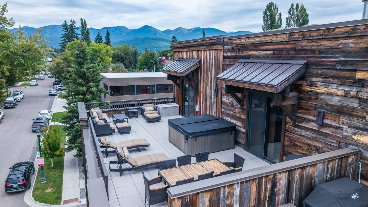 Rooftop patio with hot tub, dining table, and lounge chairs. Wood siding, mountain views, and street with cars in the background.
