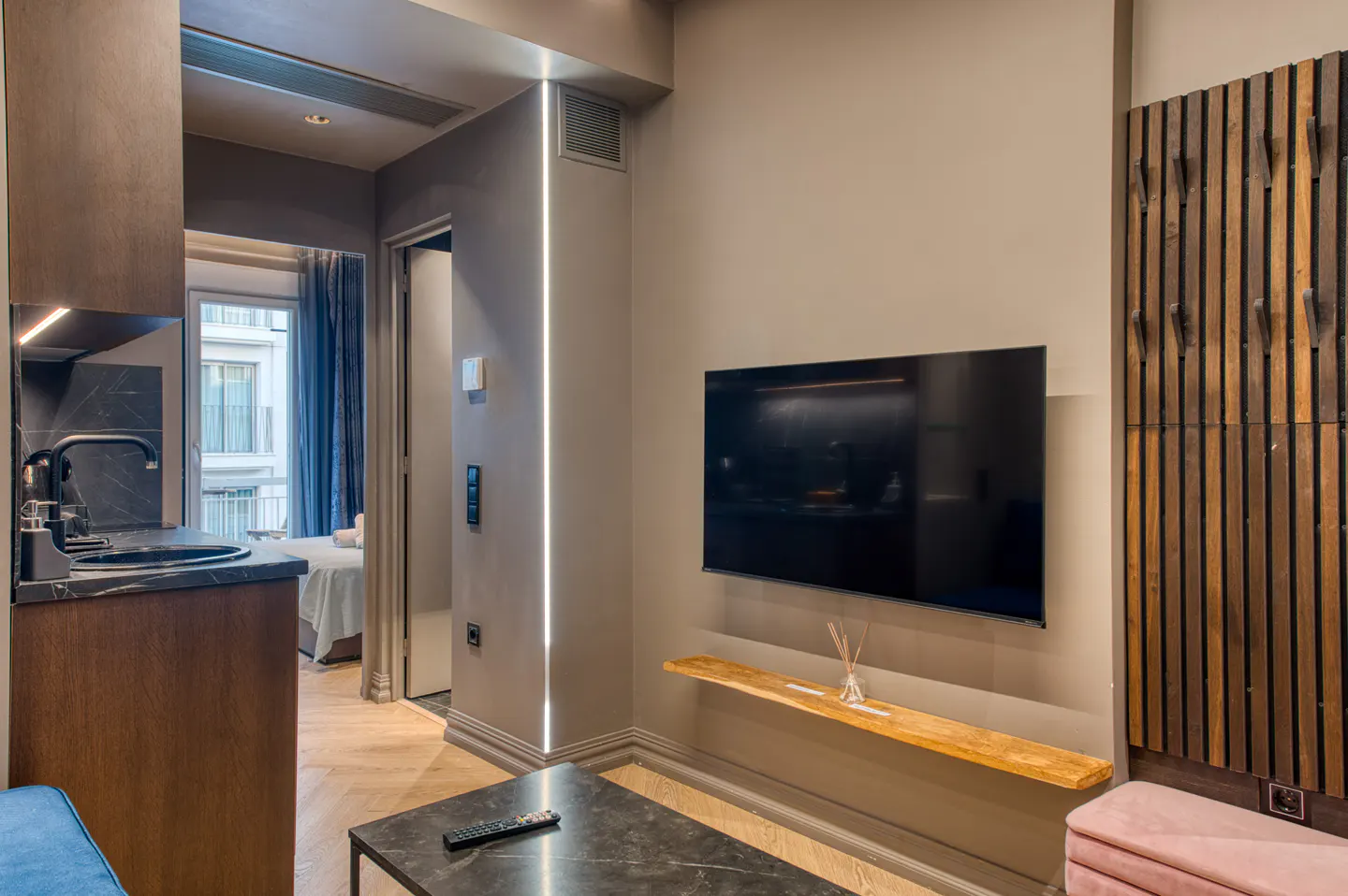 Modern apartment interior with a TV, black marble coffee table, and a small kitchen area visible in the background.