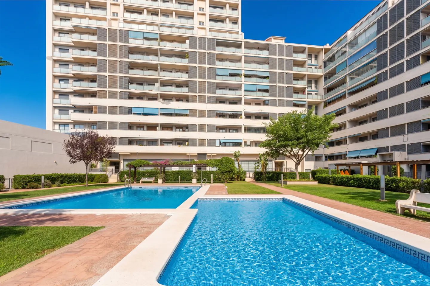 Exterior view of a tall apartment building with two blue swimming pools and green grass.