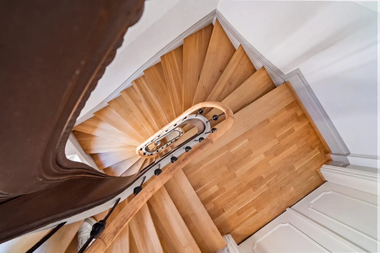 Looking down on a spiral staircase with light wood steps and a brown banister in a white room.