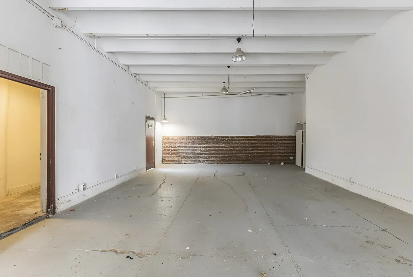 Empty commercial space with white walls, gray floor, and exposed brick accent wall. Three pendant lights hang from the white ceiling beams.