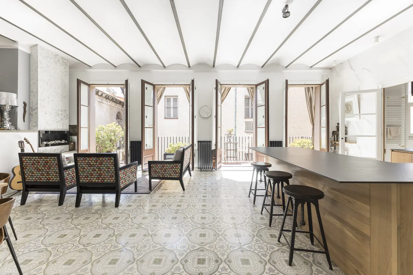 Bright living room with patterned tile floor, chairs, and open doors to a balcony. A kitchen island with stools is on the right.