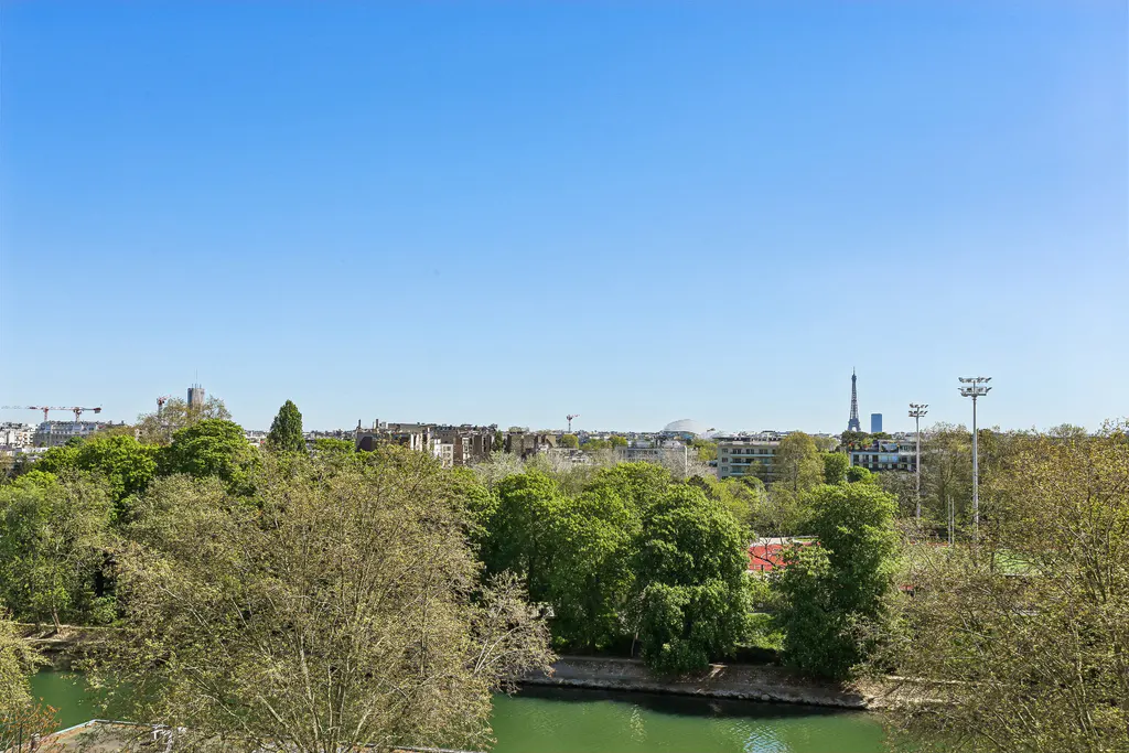 View of Paris skyline with Eiffel Tower, green trees, and river under a clear blue sky.