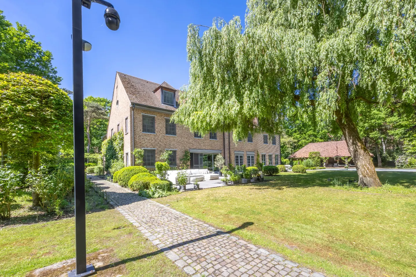 Exterior view of a large, tan brick house with a stone walkway, green lawn, and mature trees under a blue sky.