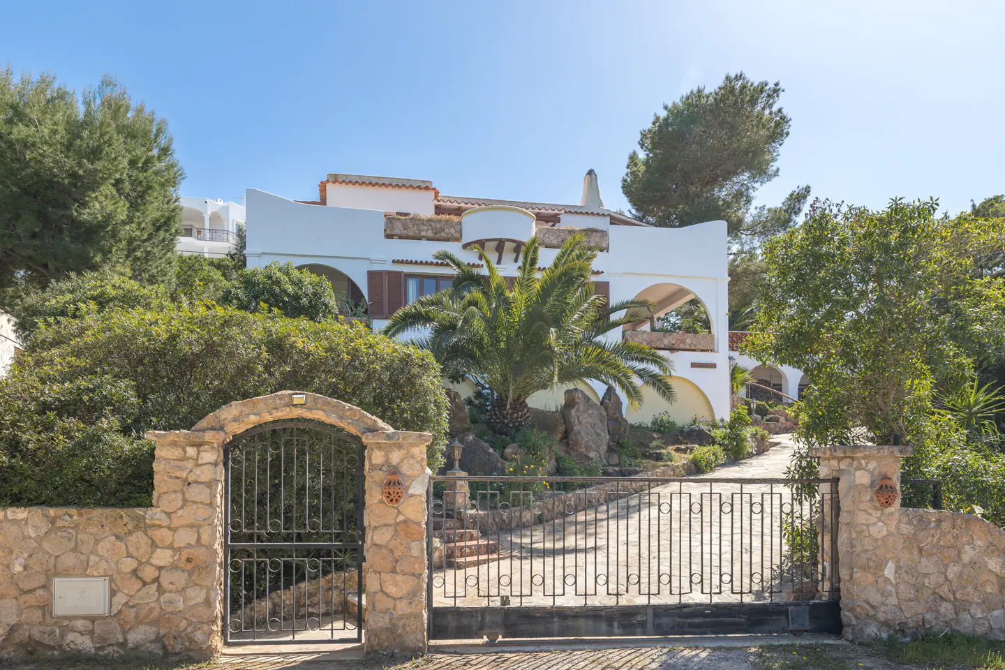 Exterior view of a white, multi-story house with arched windows, a palm tree, and a stone gate.