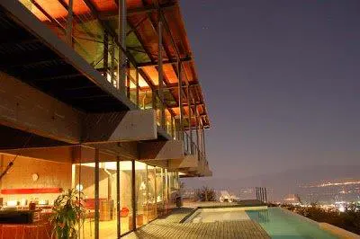 Modern home exterior at dusk with a pool. The house has large windows and a wood and metal frame. City lights are visible in the distance.