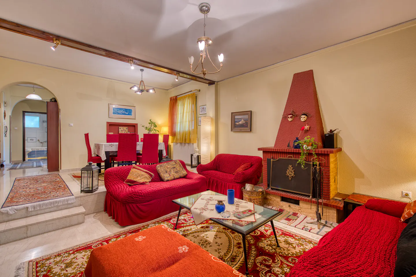 A living room with red sofas, a fireplace, and a dining area. The walls are light yellow, and the floor is tiled.
