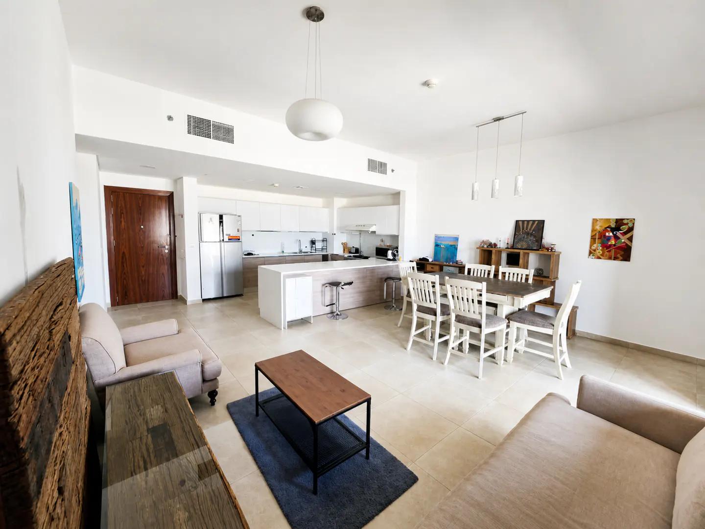 Open-concept living space with white walls, tile floors, and modern furnishings. Kitchen island, dining table, and sofas are visible.