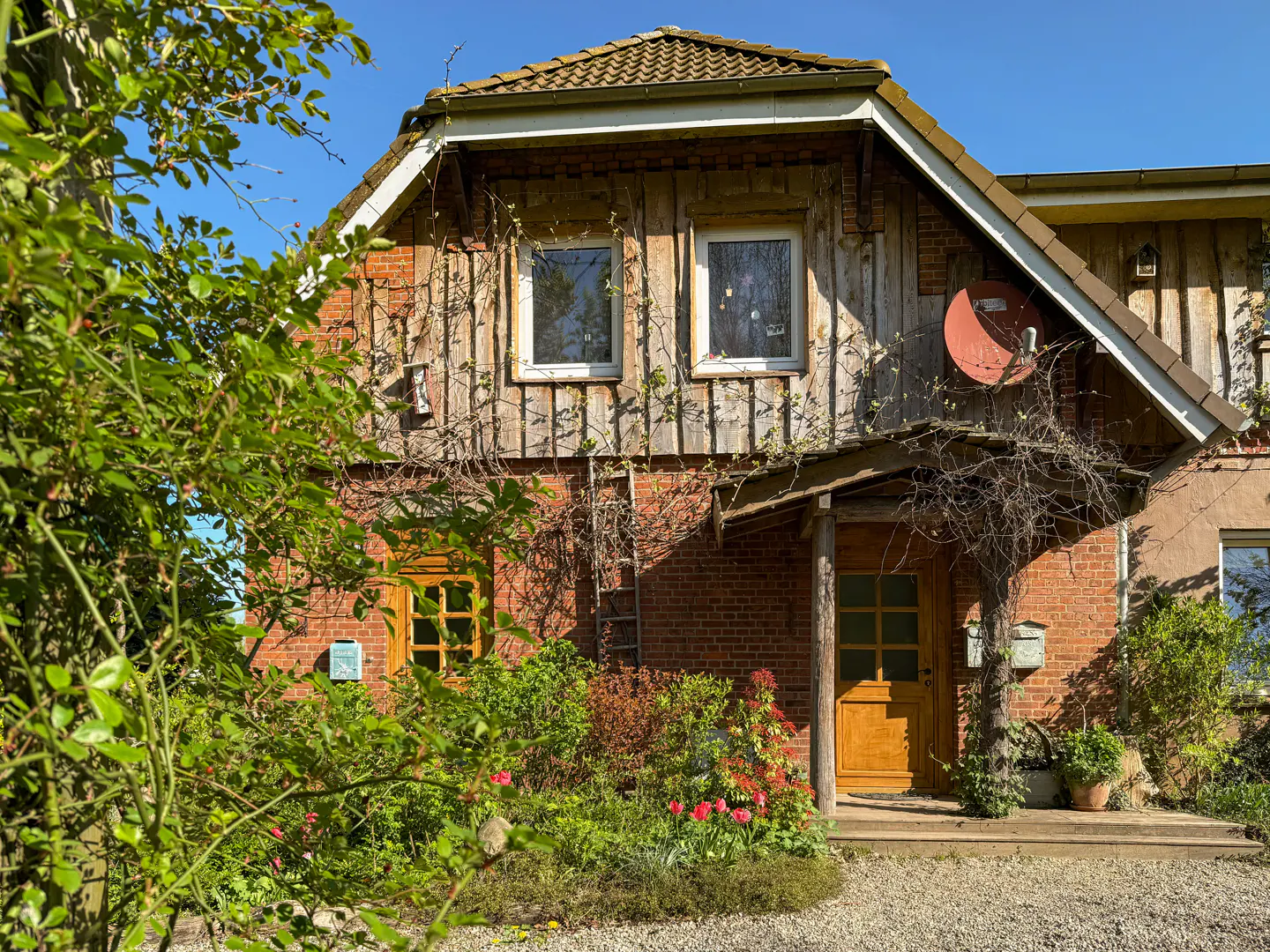 Two-story house with brick and wood siding, a brown door, and a satellite dish. Green foliage in the foreground.
