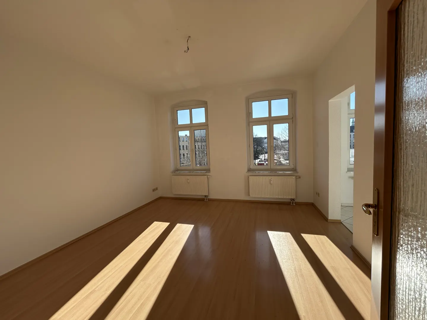Bright, empty room with wood floors, white walls, and two windows. Sunlight streams in, casting shadows. A doorway is visible on the right.